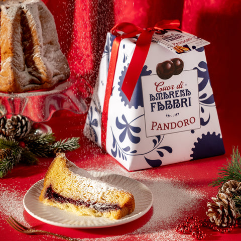 Fetta di pandoro Cuor di Amarena Fabbri su piatto, decorata con zucchero a velo. Confezione Pandoro Cuor di Amarena Fabbri sullo sfondo.