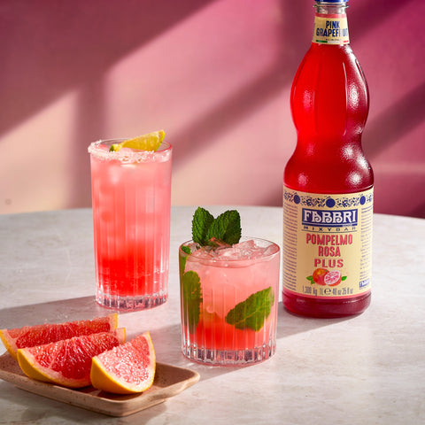 Immagine che raffigura bottiglia di Mixybar pompelmo rosa e due drink realizzati con il prodotto a fianco. Fettine di pompelmo rosa fresco in primo piano.
